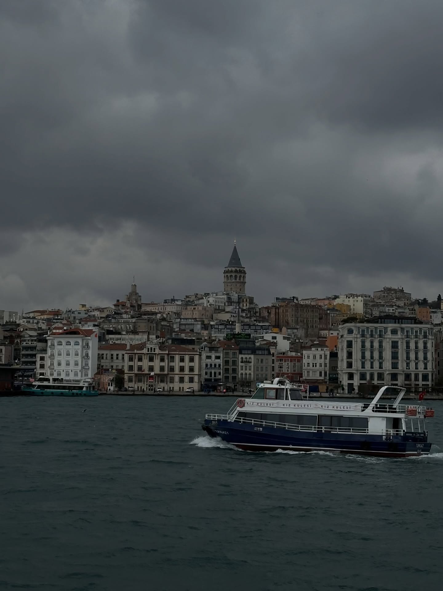 A ferry crossing the water with the Istanbul skyline and Galata Tower under storm clouds